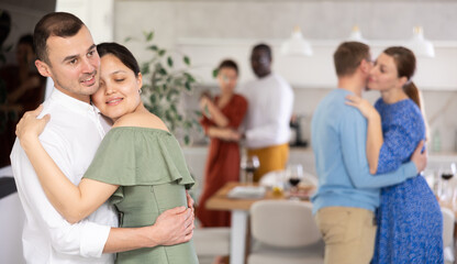 Fototapeta premium Smiling Asian woman in olive dress sharing warm slow dance with male partner, surrounded by friends at laid-back house party..