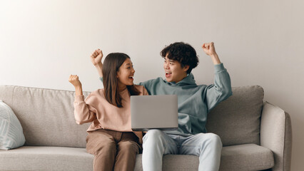 Two young adults are sitting on a couch with a laptop in front of them. They look excited and are cheering, showing signs of celebration. Their expressions show happiness.