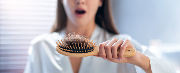 A woman stands in a bathroom holding a hairbrush with hair caught in the bristles. She looks...