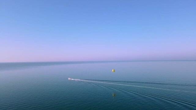 boat, parasail, parasailing, motorboat turning, sea, ocean, calm water, boat wake, yellow parachute, water sport, coastal activity, sunset horizon, pink sky, drone footage, aerial perspective, slow ci