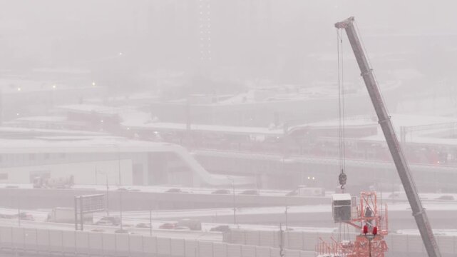 Installation of construction crane near snowy highway interchange during heavy winter snowfall, beginning of urban development and large scale infrastructure project in metropolitan area
