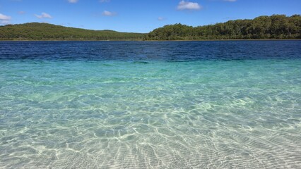 Lake McKenzie, Fraser Island, Australia - 6 January 2026. Transparent shallows over white sand fade into darker lake water, with tree-covered banks under open sky.