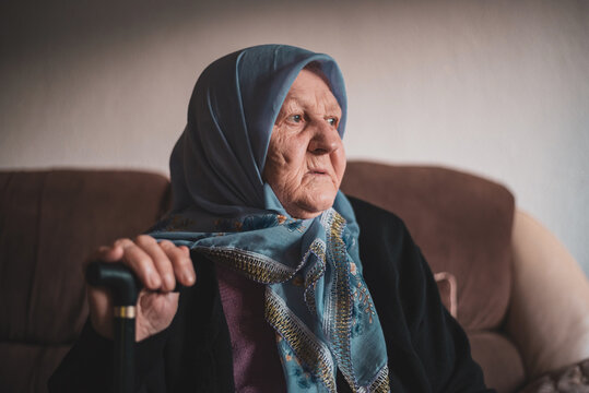 Portrait of an old thoughtful Muslim woman with her walking stick at home wearing traditional hijab, looking away.