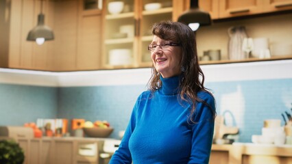 Portrait of joyful aged woman in apartment kitchen shows relaxed enjoyment during retirement. Elder person stands by the counter, embracing casual housekeeping and positive wellbeing. Camera B. © DC Studio