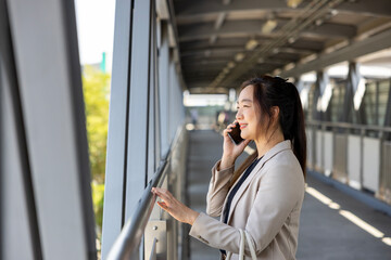 Fototapeta premium Luxury young Asian CEO woman entrepreneur using mobile phone while walking in the urban skyscraper street modern office downtown on background for business communication and urban lifestyle concept