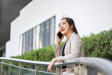 Fototapeta premium Luxury young Asian CEO woman entrepreneur using mobile phone while walking in the urban skyscraper street modern office downtown on background for business communication and urban lifestyle concept