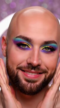 Surprised male beauty influencer with colorful rainbow eye makeup and long silver nails touching his face in a glamorous studio portrait, celebrating diversity and pride concepts.