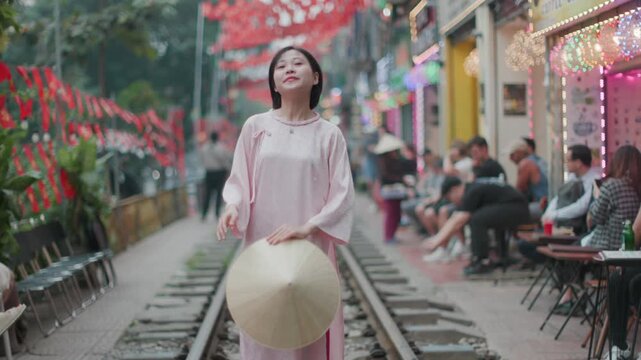 A woman in traditional dress and a conical hat poses on the train tracks in Hanoi, Vietnam. The tracks are lined with cafes and shops, creating a unique atmosphere. Train Street.