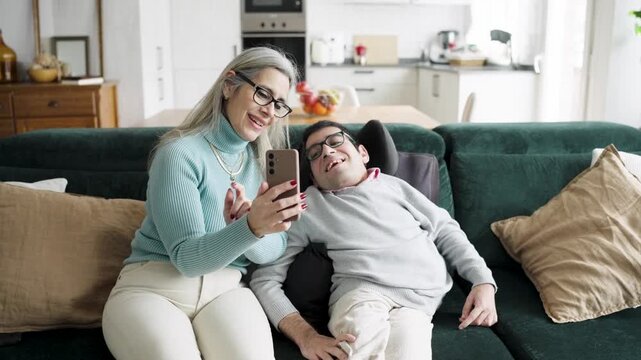 Loving mother and her son sitting together on the couch, smiling while using a smartphone for a video chat in their living room at home