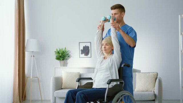Physical rehabilitation after accident. Professional man paramedic warming up hands of woman with disability sitting in wheelchair, exercising with dumbbells at home, slow motion