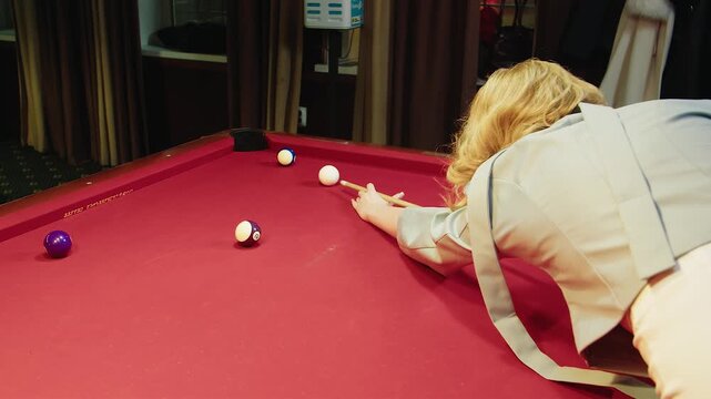 Caucasian blonde woman bending over red table practicing focused pool shot. The ball flies into the hole after hit. Multiple balls spread across felt, calm indoor club setting and evening leisure vibe