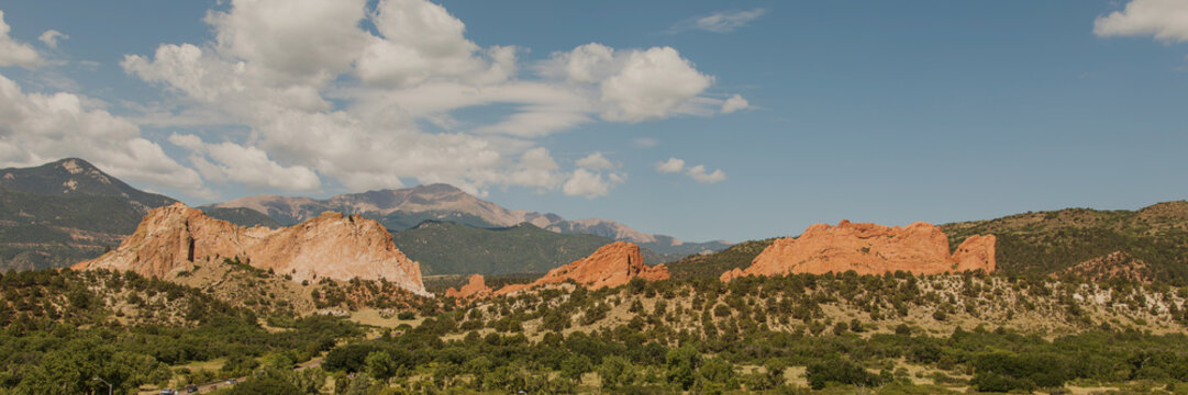 Garden of the Gods Panorama. A panoramic view showcases vibrant red rock formations against a backdrop of green foliage and mountains under a bright blue sky with scattered clouds.