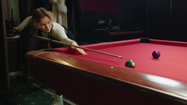 Caucasian young woman leaning for strategic club shot on red felt with determined expression stands near wooden table for billiard gaming in a club with intimate mood. A strike with cue on white ball