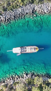 Aerial view of a wooden gulet sailing boat anchored in a clear turquoise bay surrounded by rocky coastline