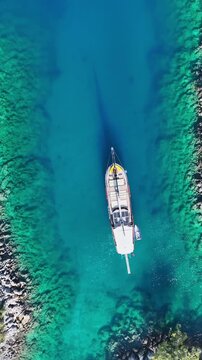 Aerial view of a traditional wooden gulet sailing through crystal clear turquoise waters near a rocky coastline