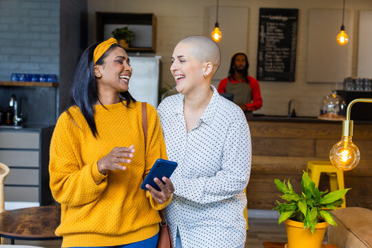 Diverse female friends laughing while holding smartphone in modern coffee shop with chalkboard menu