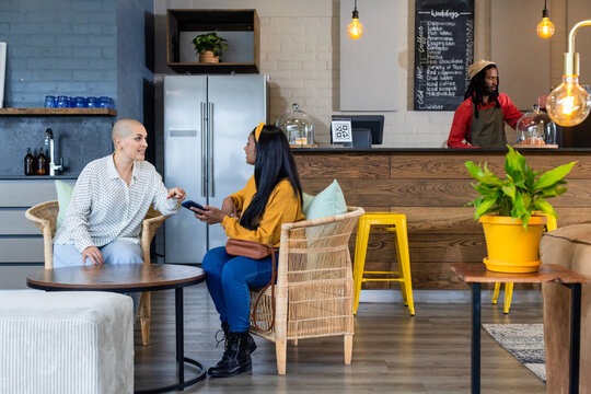 Diverse female friends chatting and using smartphone in coffee shop lounge with wicker armchairs