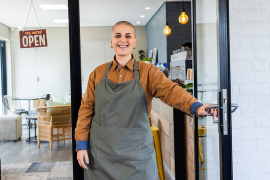 Female barista standing by door displaying Yes, we're OPEN sign in cafe with apron, coffee machine