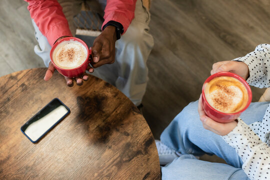 Diverse friends holding red mugs with frothy spiced coffee, checking smartphone on round cafe table