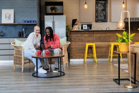 Diverse friends chatting while holding smartphone between red mugs on table in coffee shop lounge