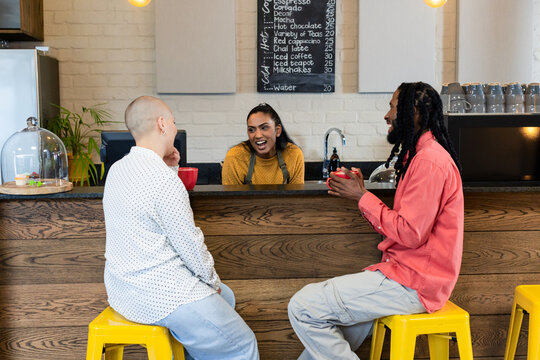 Indian barista and two customers smiling and interacting at cafe counter while holding red mugs