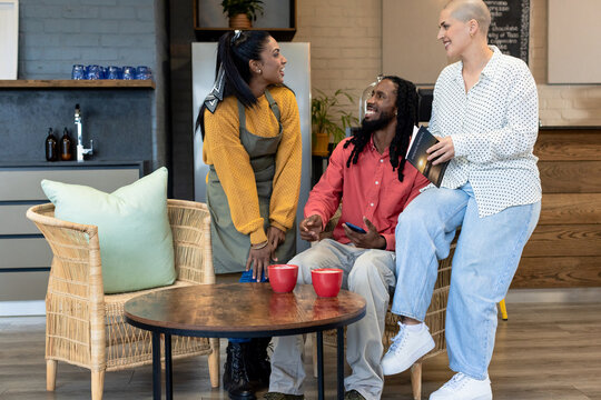 Diverse coworkers chatting around round wooden coffee table inside modern cafe lounge with red mugs