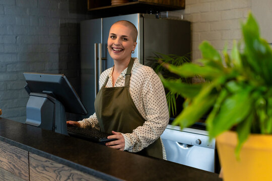 Woman standing behind counter operating terminal in cafe with fridge, dishwasher and potted plant