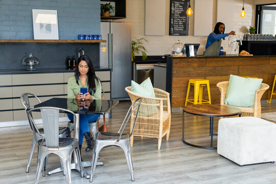 Mid adult Indian woman sitting at glass-top table in modern coffee shop lounge using smartphone