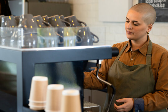Female barista wearing apron operating espresso machine at counter with takeaway cups, copy space