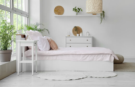 Bed with stylish pink bedding, bedside table, commode and houseplants near window in bedroom