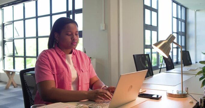 Woman in pink shirt picking up papers, referencing while typing on silver laptop, sipping water