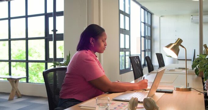 African woman wearing pink shirt triggered by charts, typing, noting data on laptop at office desk