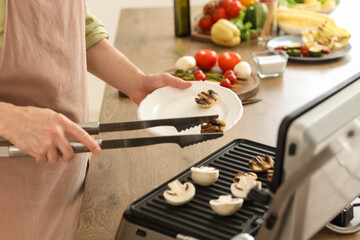 Young man putting tasty cooked mushrooms on plate from modern electric grill in kitchen, closeup
