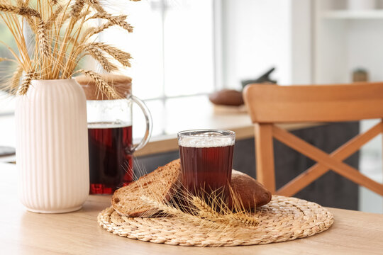 Fresh kvass in glass with rye bread and wheat ears on wooden kitchen table, rustic homemade fermented drink in cozy bright interior