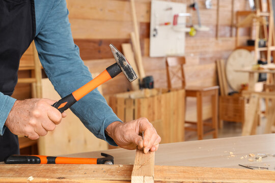 Mature carpenter hammering nail into wooden plank at table in shop, closeup