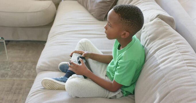 African American boy green tee starting game, holding pad, trying to win on sofa, copy space