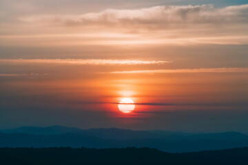 Obraz premium Beautiful sunset over distant mountain layers with warm orange sky and soft clouds, creating a peaceful natural background and serene landscape atmosphere.