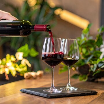 Pouring red wine into two elegant glasses on a wooden table with a blurred background.
