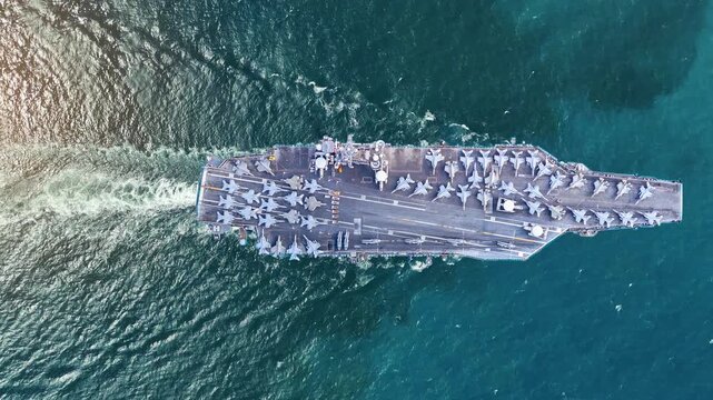 nuclear navy aircraft carrier sailing in the deep blue ocean. Cinematic shot of a military warship deck loaded with fighter jets leaving a white wake.