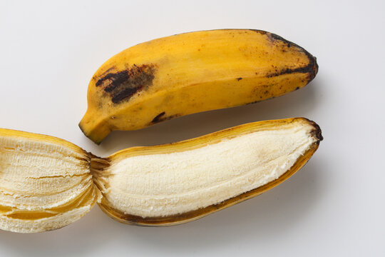 Closeup of two saba bananas (pisang kepok) on a white background, one fully intact and the other partially peeled to reveal the fruit inside.