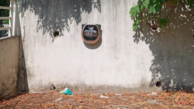 A robotic vacuum cleaner tries to climb a dilapidated concrete wall in the shade of trees, in an open area where fallen leaves are scattered on the ground, and falls down.
