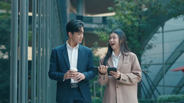 Two young Asian business colleagues walking and discussing project or having a meeting with a digital tablet outside a modern office building. Concept of teamwork, partnership, and professional lifest