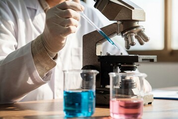 Fototapeta premium Scientist gloved hand with pipette next to microscope and chemical beakers