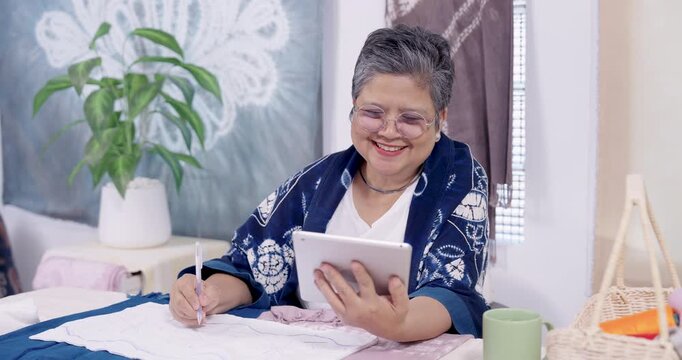 Senior Asian woman smiles, holds tablet, compares shibori template with cloth, then draws guide line for resist stitching, preparing handmade indigo dye textile in home studio workshop business.