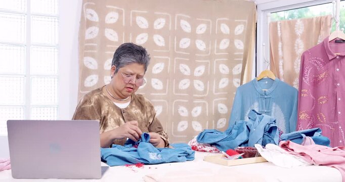 Senior Asian woman artisan sits near laptop, unpicks red resist stitching from blue cotton shirt, freeing shibori pattern before indigo dyeing, running handmade textile small business in home studio.