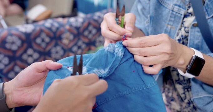 Hands pull red thread while helper steadies blue cotton shirt, removing resist stitching loops to reveal shibori tie-dye pattern before indigo dyeing, small business craft process in home studio.
