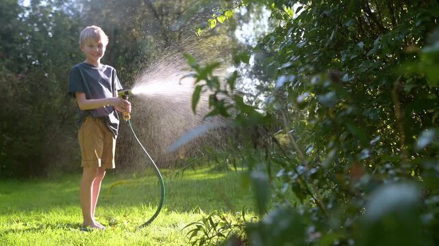Funny boy watering the bushes and playing with garden hose with sprinkler in sunny backyard on hot summer day. Happy child having fun with water splashes. Summer outdoor activity for kids.