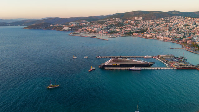 Kusadasi, Turkey. A luxury mega yacht docked at the port in the twilight after sunset. Aerial view of the vessel with dark hull against the city and glowing evening sky. Aerial View