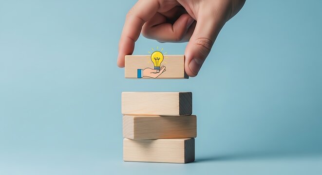 Hand placing wooden block with lightbulb on of a stack of wooden blocks against blue background