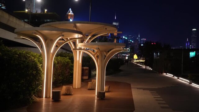 Night pedestrian area with illuminated architectural canopies. A lit walkway with urban landscaping elements and greenery along the path. 